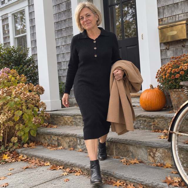 Nancy in the black Elyon dress with ankle boots, a camel coat over her arm